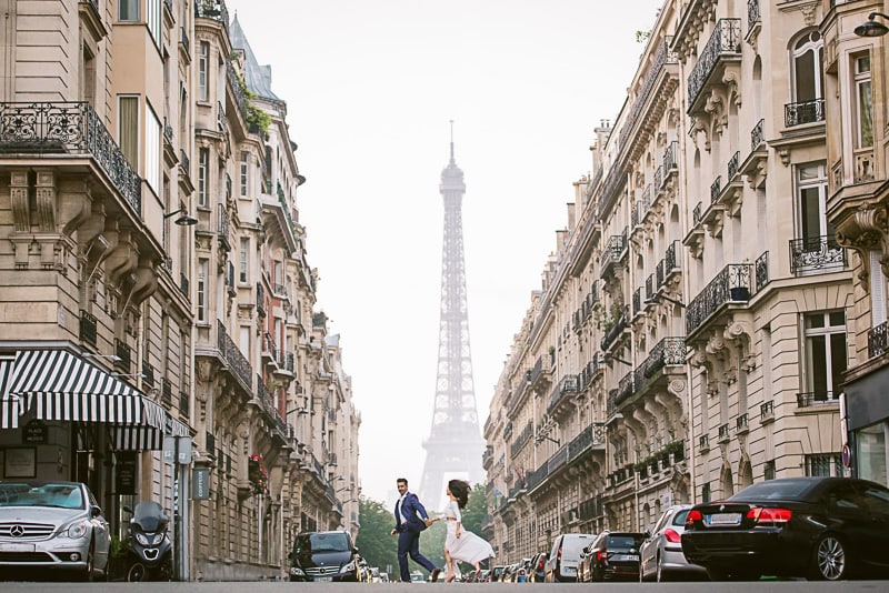 Newly engaged couple running across the street between two buildings and the Eiffel Tower in the background